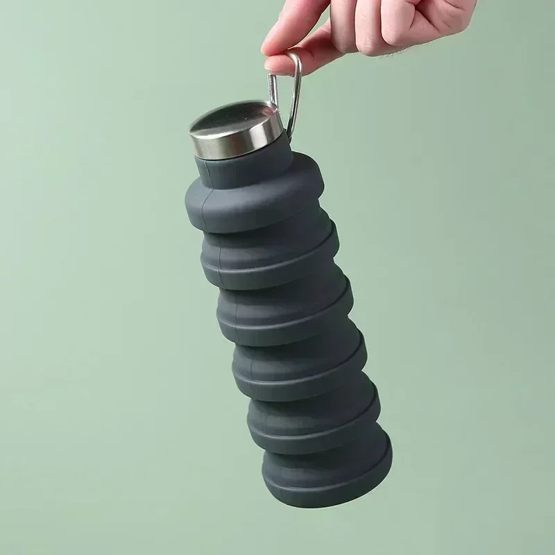 Hand holding a charcoal Squishly collapsible silicone water bottle by its carabiner clip against a sage green background