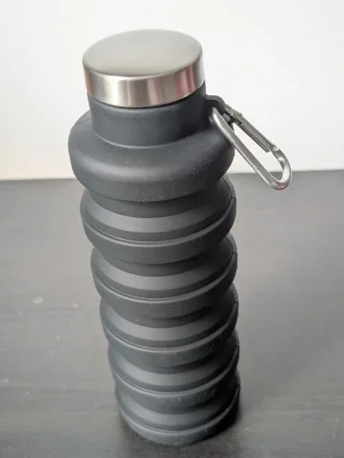 Charcoal-grey collapsible silicone water bottle standing fully expanded, stainless steel cap and carabiner clip on a dark surface — photo from a verified buyer