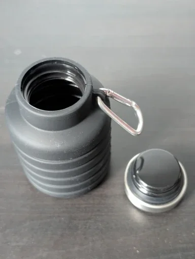 Dark grey collapsible water bottle with its stainless steel cap unscrewed and set beside it, showing the wide drinking mouth and carabiner — photo from a verified buyer