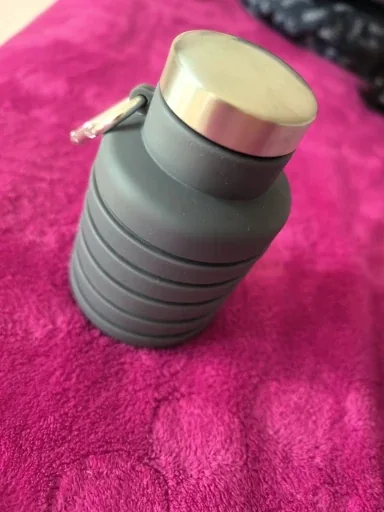 Grey-green collapsible silicone water bottle folded down small with a metallic screw cap, resting on a pink blanket — photo from a verified buyer