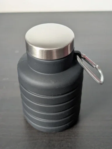 Dark grey collapsible water bottle folded down to a compact size with its stainless steel cap and carabiner, on a dark surface — photo from a verified buyer